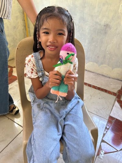A child wearing dungarees, sitting in a chair, holding a knitted doll and smiling